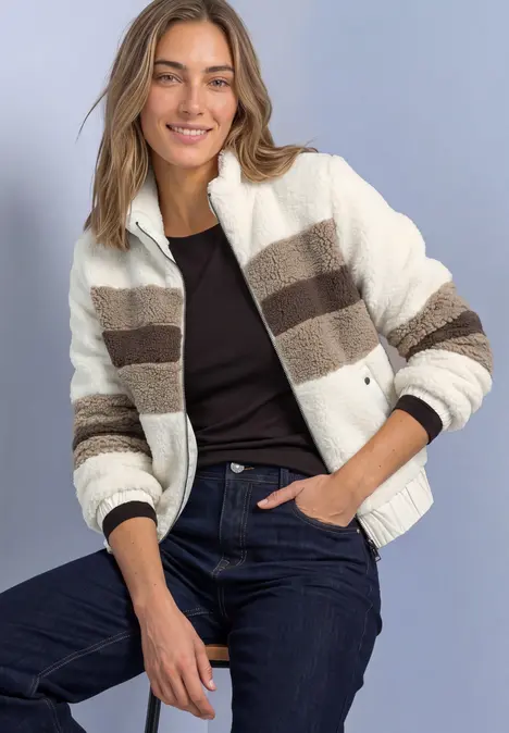 Frau sitzt lächelnd, trägt gestreifte, flauschige Fleecejacke in Creme- und Brauntönen, schwarzen Top und Jeans.