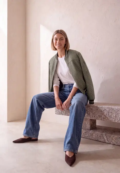 Frau sitzt lässig auf einer Steinbank, trägt Jeans, weißes Shirt und grünes Sakko, lächelt in minimalistischem Raum.