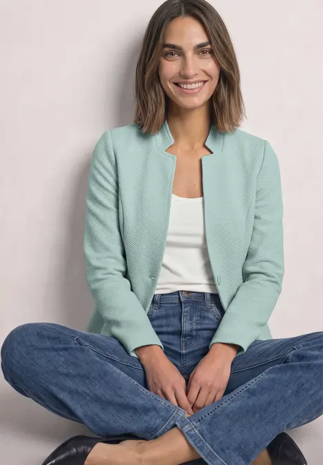 Eine lächelnde Frau sitzt im Schneidersitz und trägt ein hellgrünes Sakko, weißes Top und blaue Jeans.