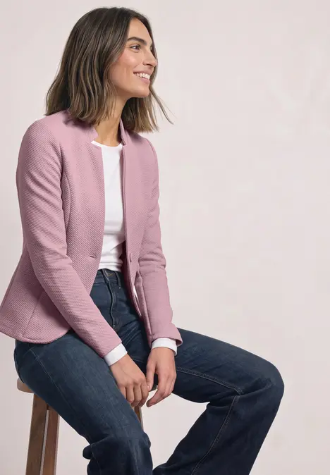 Frau auf Hocker, rosa Blazer, weißes Shirt und Jeans, lächelt und blickt seitlich vor hellem Hintergrund.