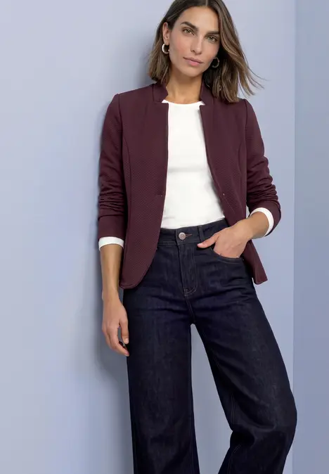 Frau mit burgunderfarbenem Blazer, weißem Shirt und weit geschnittenen Jeans vor schlichtem Hintergrund.