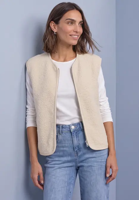 Eine Frau in weißem Langarmshirt, beigefarbener Fleeceweste und Jeans vor hellblauem Hintergrund.