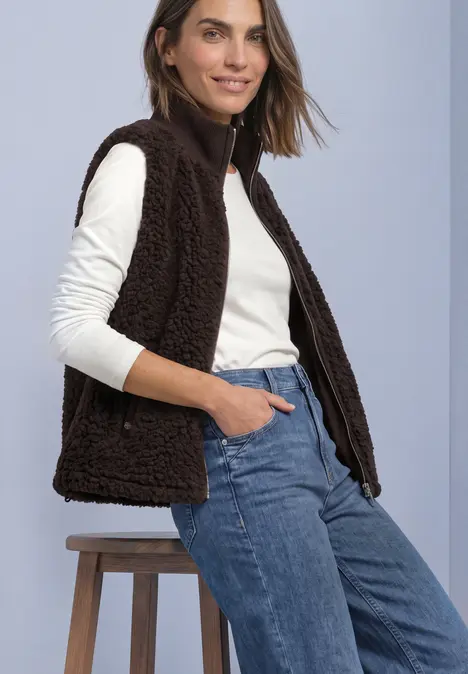 Femme portant un top blanc à manches longues, une veste marron pelucheuse et un jean bleu, assise sur un tabouret.