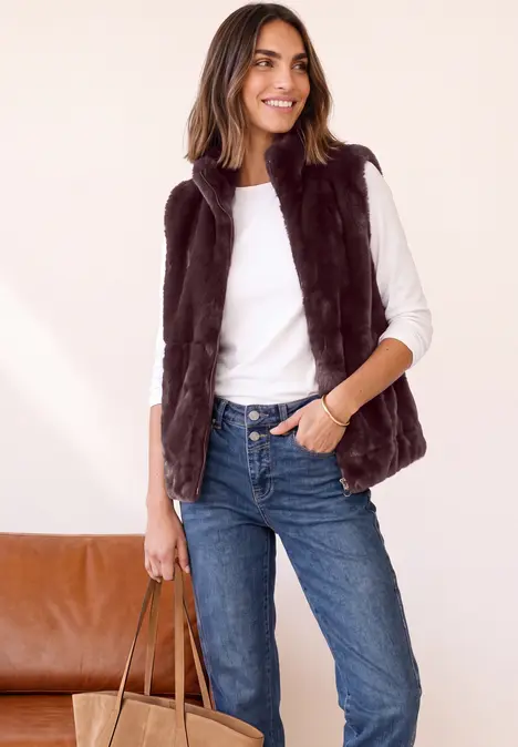 Eine lächelnde Frau trägt weiße Bluse, braune Weste, blaue Jeans und hält eine Tasche.