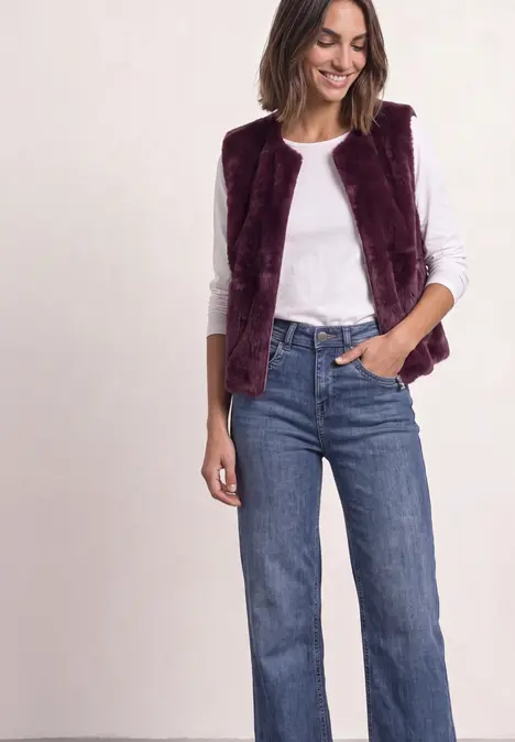 Femme portant un t-shirt blanc, un gilet bordeaux en fausse fourrure et un jean bleu large.