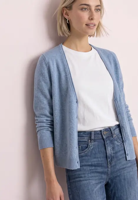 Frau in hellblauem Cardigan, weißem T-Shirt und blauen Jeans vor hellrosa Hintergrund.