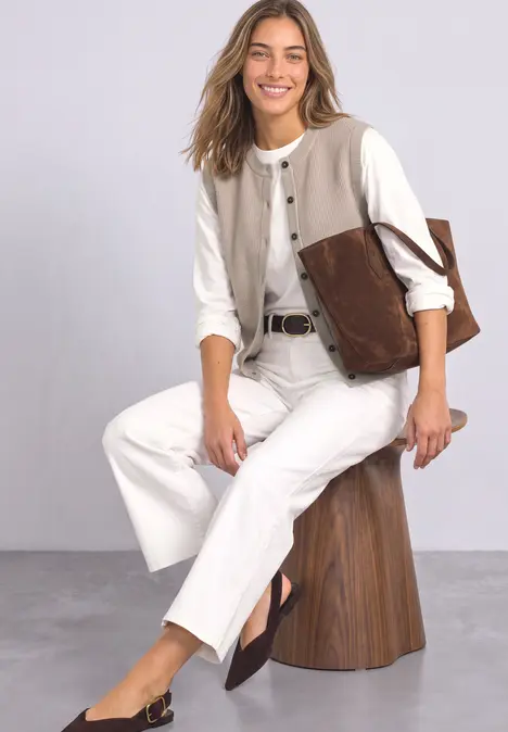 Eine Frau sitzt auf einem Hocker, trägt weiße Hose, weiße Bluse und braune Accessoires, lächelt freundlich.