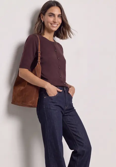 Eine Frau in braunem Cardigan und dunklen Jeans mit brauner Schultertasche lächelt vor neutralem Hintergrund.