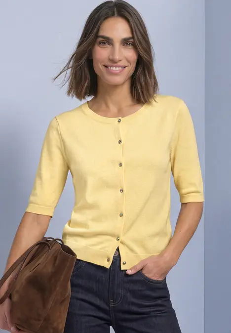 Frau mit gelbem, knöpfbarem Kurzarm-Pullover, dunklen Jeans und brauner Umhängetasche.