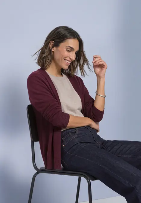 Eine lächelnde Frau sitzt auf einem Stuhl, trägt einen weinroten Cardigan, beige Top und blaue Jeans.