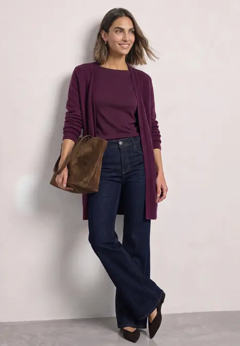 Frau trägt dunkelrotes Oberteil mit Cardigan, weite blaue Jeans, schwarze Schuhe und braune Wildledertasche.
