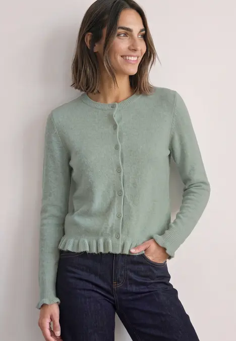 Vrouw met halflang haar draagt groen vest met knopen en ruches aan de zoom, glimlacht zacht.