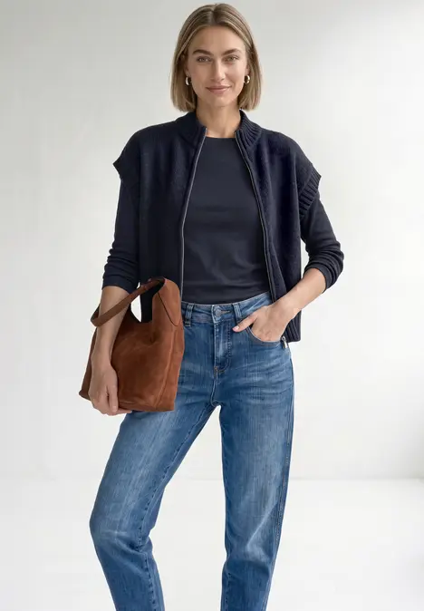 Eine lächelnde Frau trägt blaue Jeans, ein dunkelblaues Shirt, eine Bomberjacke und eine braune Tasche.