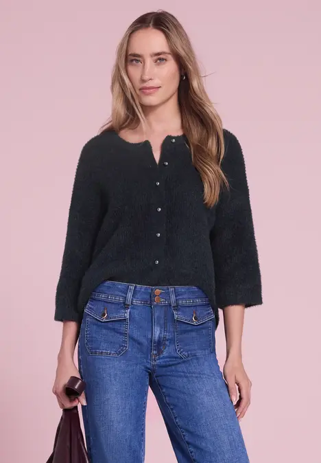 Een vrouw in een zwart vest met knopen en blauwe jeans voor een roze achtergrond.