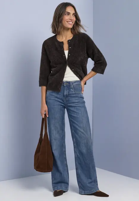 Frau vor blauem Hintergrund, in weitem Jeans, weißem Shirt und schwarzer Jacke, trägt braune Tasche.