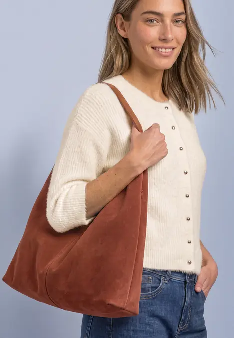 Eine Frau trägt lächelnd eine braune Wildleder-Hobo-Tasche an der Schulter, dazu cremefarbener Cardigan und Jeans.
