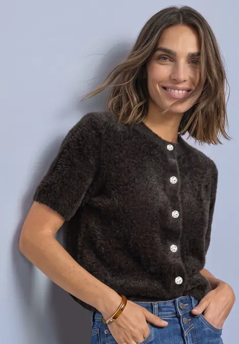 Eine lächelnde Frau trägt einen kurzärmligen, flauschigen, dunklen Cardigan mit weißen Knöpfen, Hände in den Taschen.