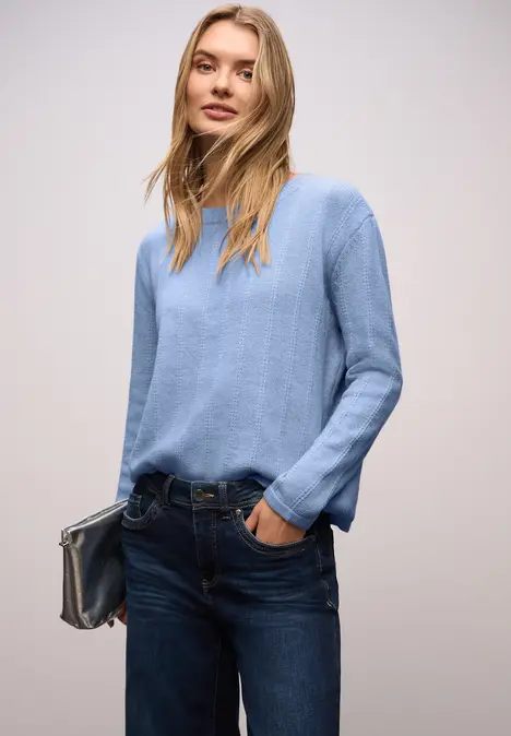 Eine Frau in hellblauem Pullover, dunkelblaue Jeans, hält eine Clutch und steckt die Hand lässig in die Tasche.