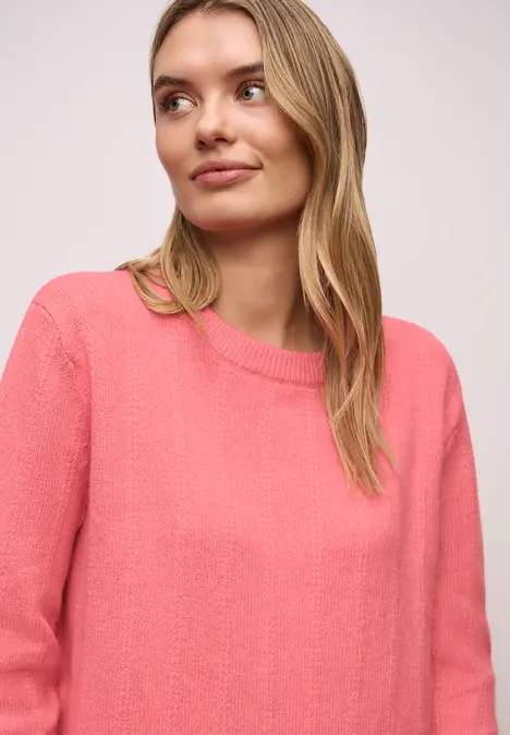 Frau mit langem blonden Haar in lockerem pinken Pullover vor hellem Hintergrund.