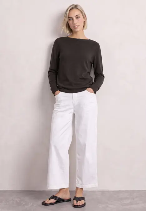 Frau in schwarzem Pullover, weißer weiter Hose und schwarzen Sandalen vor neutralem Hintergrund.
