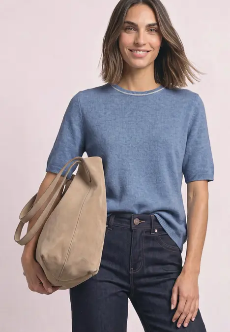 Eine Frau im blauen kurzärmligen Pullover und dunklen Jeans trägt lächelnd eine große beige Tasche.
