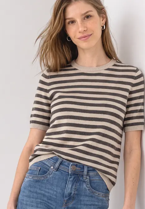Junge Frau in gestreiftem Kurzarmshirt und Jeans lehnt lässig an einer Wand.