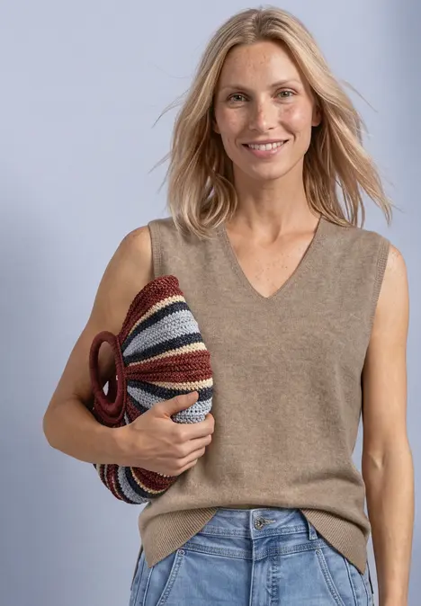 Frau mit blondem Haar, trägt einen ärmellosen Pullover, Jeans und hält eine farbige gestreifte Tasche.
