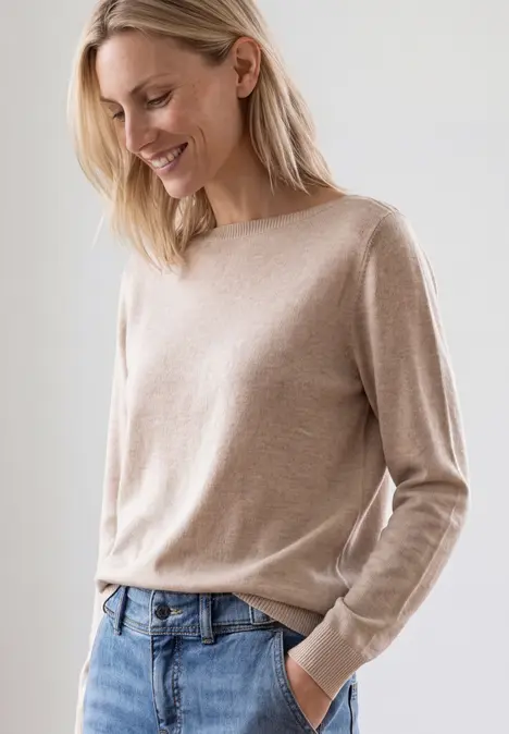 Eine lächelnde Frau trägt einen beigen Pullover und Jeans vor schlichtem Hintergrund.