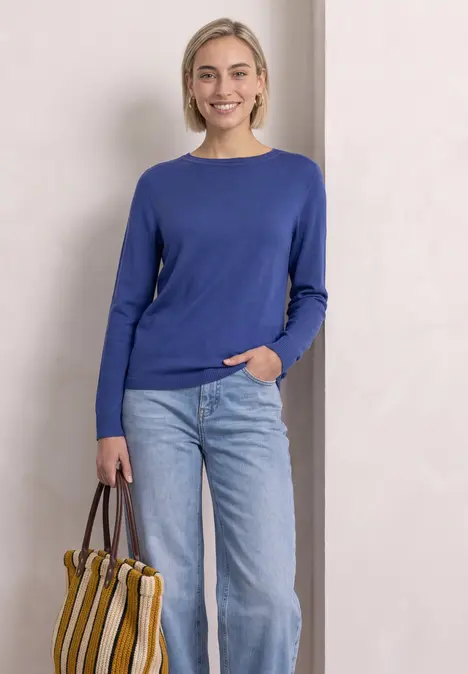 Eine Frau steht vor hellem Hintergrund und trägt einen blauen Pullover, Jeans und eine gestreifte Tasche.