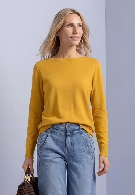 Eine Frau mit senfgelbem Pullover und blauen Jeans hält eine Handtasche und blickt seitlich.