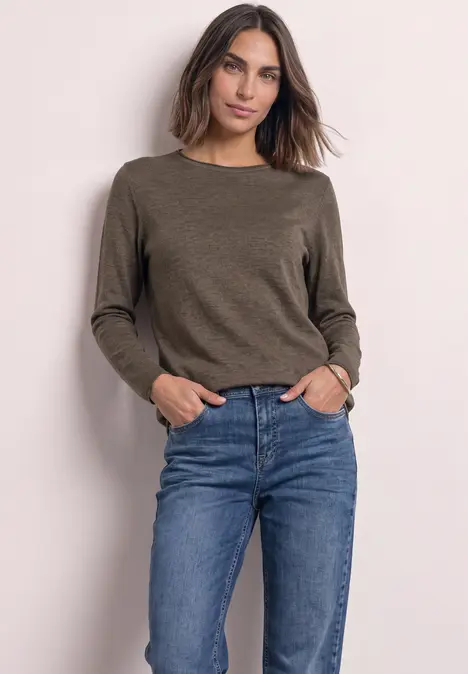 Vrouw in olijfgroene trui en blauwe jeans voor een lichte achtergrond.