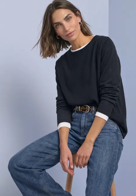 Vrouw zit op kruk, draagt zwarte trui met witte kraag en blauwe jeans, voor een grijze achtergrond.