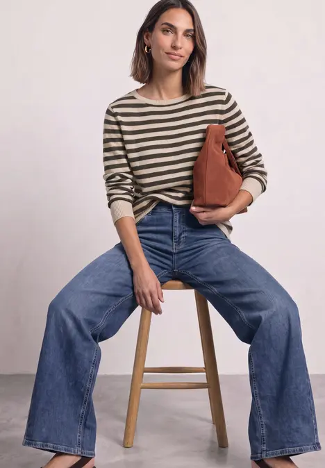 Frau sitzt auf Hocker, trägt gestreiften Pullover, weite Jeans und hält eine braune Tasche.