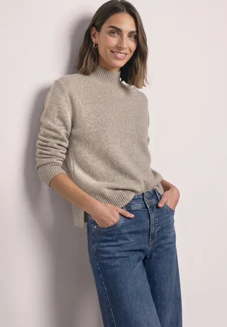 Eine lächelnde Frau in beigem Pullover und blauen Jeans lehnt lässig mit den Händen in den Taschen.