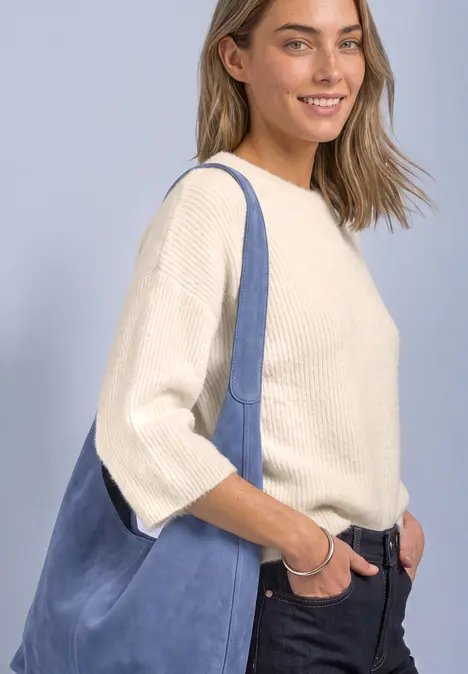 Junge Frau mit cremefarbenem Pullover, Jeans und großer blauer Umhängetasche, die lächelt.