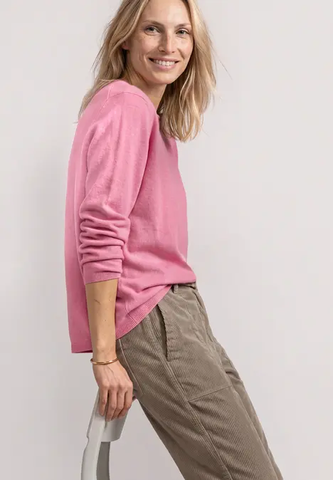 Frau im rosa Pullover und beigefarbenen Hosen, seitlich stehend, lächelnd vor hellem Hintergrund.