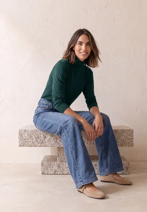 Eine Frau sitzt auf einer Steinbank, trägt grünen Rollkragenpullover, Jeans und Ballerinas.