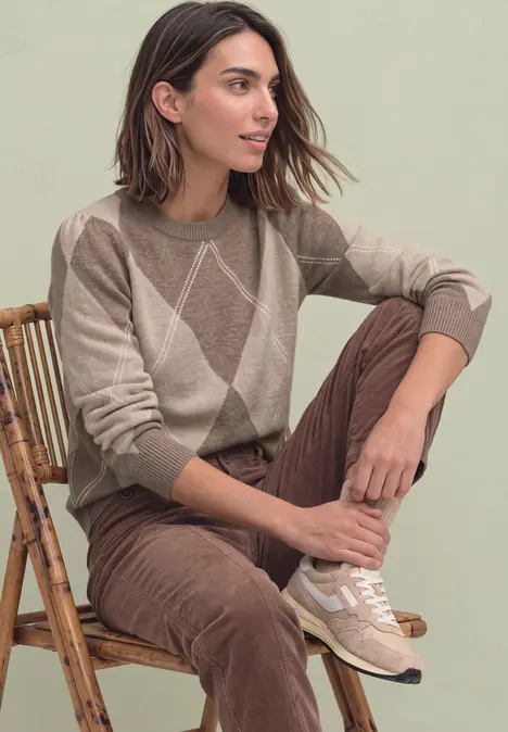 Eine Frau sitzt auf einem Rattanstuhl, trägt einen beigen Pullover mit Rautenmuster und braune Cordhose.
