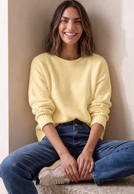 Une femme souriante, assise en tailleur, porte un pull jaune et un jean bleu devant un mur beige.
