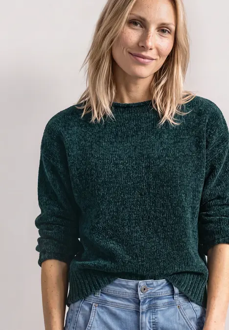 Femme blonde en pull vert et jean, souriante, sur fond clair.