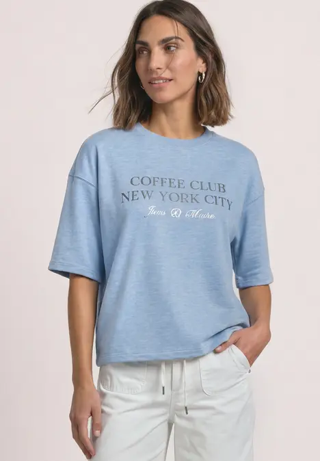 Frau in hellblauem T-Shirt mit Aufschrift 'COFFEE CLUB NEW YORK CITY' und weißer Hose, lächelnd.