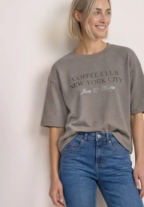 Frau trägt graues T-Shirt mit Aufschrift 'Coffee Club New York City' und blaue Jeans.