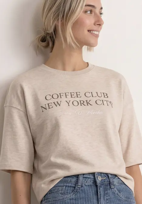 Frau in beigem „Coffee Club New York City“-T-Shirt und Jeans, lächelt seitlich.
