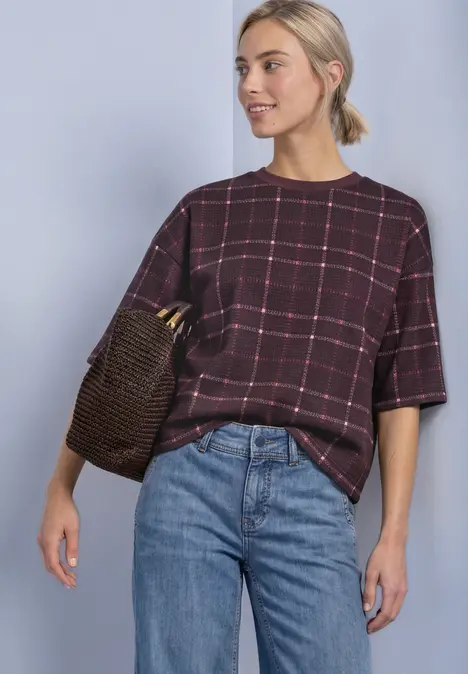 Frau trägt karierte, bordeauxrote Bluse mit halblangen Ärmeln, Jeans und geflochtene Tasche.