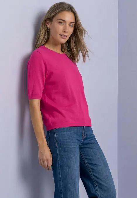 Eine Frau steht seitlich in einem pinken Kurzarmshirt und blauen Jeans.