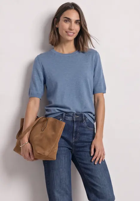 Eine Frau trägt ein blaues Kurzarm-Shirt und Jeans, hält eine braune Tasche in der Hand.