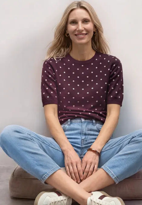 Blonde Frau im Schneidersitz vor weißer Wand, trägt gepunktetes lila Shirt und blaue Jeans.