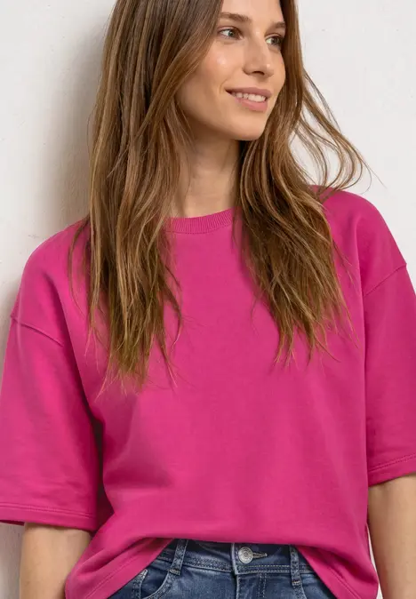Junge Frau mit locker sitzendem pinkfarbenem T-Shirt lächelt seitlich.