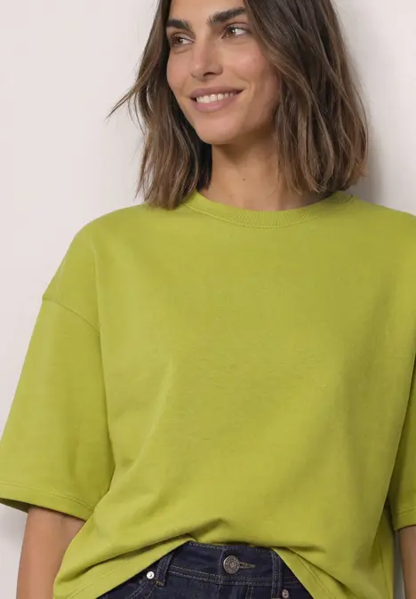 Eine lächelnde Frau trägt ein lockeres, kurzärmeliges, hellgrünes Shirt.