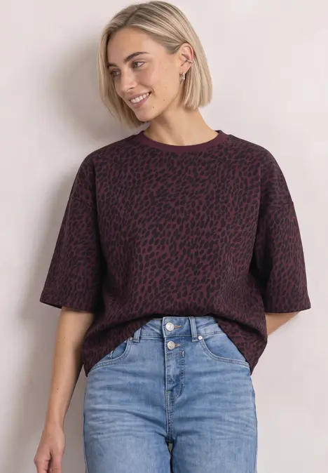 Junge Frau mit blondem Bob, violett-gemustertem Kurzarm-Pullover und hoch taillierter Jeans vor hellem Hintergrund.